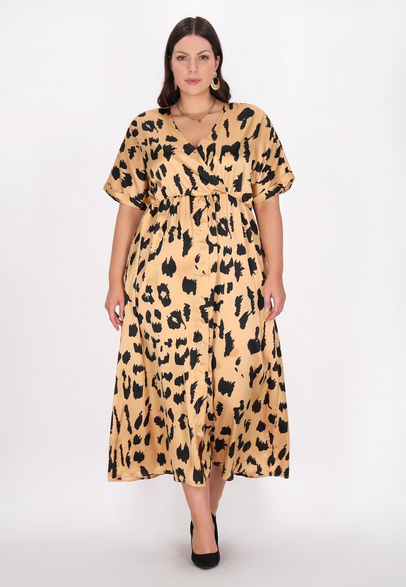 Beige leopardmønstret maxikjole med indpakningsdesign, korte ærmer, en indsnøret talje og en glat, skinnende tekstur. Sorte højhælede sko bemærket.