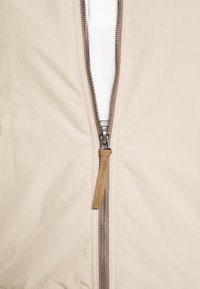 Gros plan d'une fermeture éclair beige d'une veste partiellement ouverte montrant un tissu blanc en dessous, avec une languette en tissu marron sur le curseur de la fermeture.