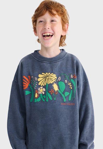 Niño pelirrojo con pecas sonriendo y vistiendo una sudadera azul con ilustraciones coloridas de flores en el pecho.