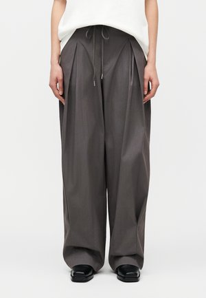 Persoană care poartă pantaloni gri largi, cu picioare largi, pliuri în față și talie cu șnur, asortați cu pantofi negri și un top alb.