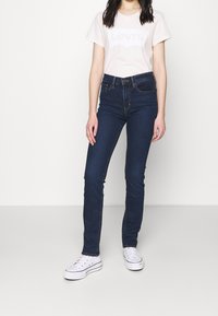 Tmavě modré džíny skinny v kombinaci se světle růžovým tričkem s logem Levi's. Boty jsou bílé tenisky s černými akcenty.