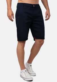 Shorts navy realizzati in misto cotone, con orli arrotolati con accenti rossi, tasche frontali e chiusura con bottone. Abbinati a sneakers bianche.