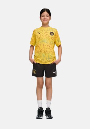 Enfant debout les mains dans les poches, portant un maillot de sport jaune à motif, un short noir, des chaussettes blanches et des baskets Puma noires.