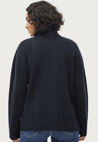 Marineblauwe gebreide coltrui met ribbelstructuur, gevallen schouders en ontspannen pasvorm. Draagt comfortabel met jeans, waarbij de details op de rug goed zichtbaar zijn.