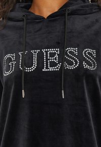 Felpa nera in velluto con un audace logo "GUESS" in strass, cordini regolabili e texture liscia.