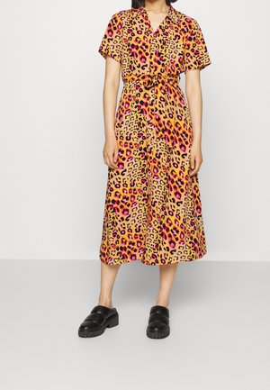 Kvinde iført en kortærmet kjole i midterlængde med en lyserød, orange og sort leopardprint, kombineret med sorte slip-on sko.