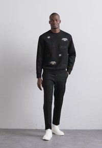 Črna majica s kapuco z teksturiranimi pravokotniki in grafikami, v kombinaciji z krojim črnimi hlačami in belimi supergami, na nevtralni podlagi.