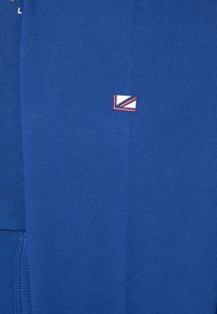 Blaues Sweatshirt aus strukturiertem Stoff. Verfügt über ein kleines Logo in Rot und Weiß, das sich auf der Vorderseite, nahe der unteren linken Ecke, befindet.