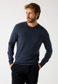 Uomo con barba che indossa un maglione a trama blu navy e pantaloni neri, con le mani in tasca, che guarda a sinistra su uno sfondo bianco uniforme.