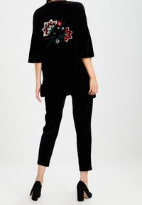 Femme portant un pantalon en velours noir et un haut en velours noir ample avec une broderie florale colorée dans le dos, associé à des talons hauts noirs.