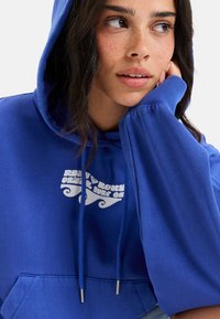 Sudadera azul con capucha, que presenta un logotipo gráfico blanco. Confeccionada en una tela suave, tiene un bolsillo frontal y mangas anchas.