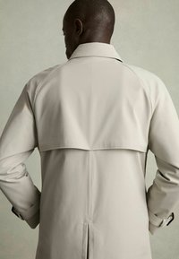 Beež trench coat laiaga kraega, kokkuvolditud flapi detailiga, sobitatud varrukatega ja tagant ventillatsiooniga. Sile materjal sujuva viimistlusega.
