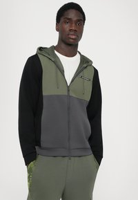 Sweatshirt com capuz em verde oliva e preto, apresentando um fecho de zíper, design em blocos de cor e detalhe do logotipo no peito. Textura de tecido suave.