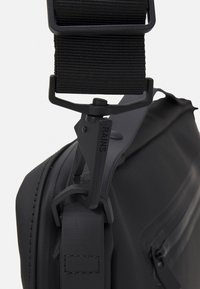 Schwarze wasserdichte Tasche mit glatter, matter Oberfläche. Verfügt über einen robusten Tragegurt und eine verstellbare Schnalle mit "RAINS"-Markierung. Reißverschlussdetails sichtbar.