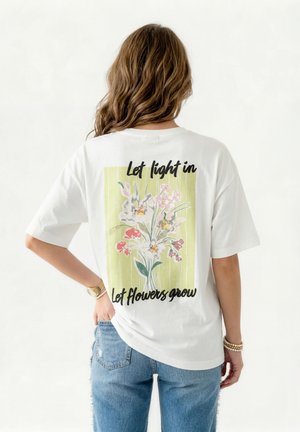 Γυναίκα που φοράει λευκό T-shirt με πολύχρωμο floral σχέδιο και το κείμενο «Άσε το φως να μπει, άσε τα λουλούδια να μεγαλώσουν» στην πλάτη, συνδυασμένο με μπλε τζιν.