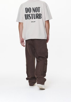 T-shirt grigio chiaro con stampa "NON DISTURBARE" sul retro, abbinato a pantaloni cargo larghi di un marrone scuro con tasche laterali.