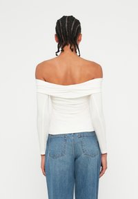 Off-the-shoulder white top with long sleeves, featuring gathered detailing along the sides. Paired with high-waisted blue jeans.