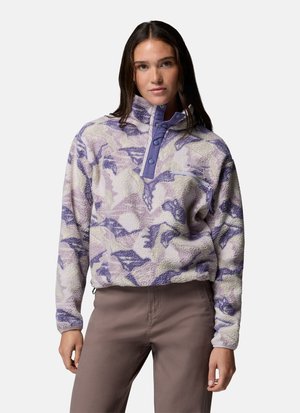 Frau trägt einen lila-beigen gemusterten Fleece-Pullover mit Druckknöpfen und Reißverschlusstasche auf der Brust, kombiniert mit braunen Hosen.