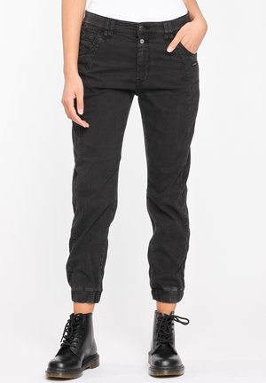 Femme portant un pantalon jogger noir taille haute avec des poignets élastiques et des bottes de combat noires à lacets, mains reposant de manière décontractée.