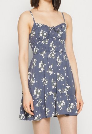 Robe fleurie avec un fond bleu foncé, motif de fleurs blanches et vertes, bretelles réglables, taille froncée et ourlet à volants.