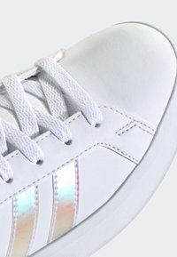 Bijela tenisica s gornjim dijelom od teksturirane kože, tradicionalnim vezicama i tri iridescentne pruge. Glatki gumeni potplat s detaljima šavova.