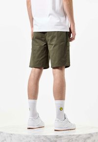 Shorts vert olive avec une coupe décontractée, ourlets repliés, poches latérales, accompagnés de chaussettes blanches avec un smiley jaune et de baskets blanches.