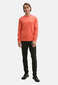 Pulover coral cu guler rotund, cu manșete și tiv din tricot, purtat cu pantaloni negri, ajustați și pantofi negri cu șireturi.