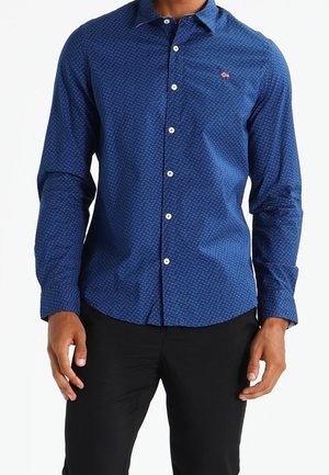 Homme portant une chemise ajustée à manches longues bleu foncé à motifs et un pantalon noir, debout, les mains détendues le long du corps.