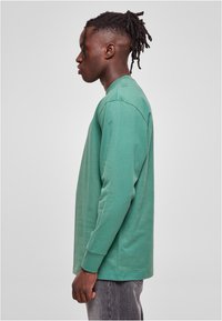 Camiseta de manga larga verde hecha de algodón, con escote redondo y corte recto, mostrada en un modelo de perfil.