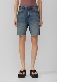Dżinsowe szorty z wysokim stanem, postrzępionym wykończeniem i klasycznym pięciopocketowym krojem w średnim odcieniu niebieskiego. Połączone z czarnymi sandałami na platformie.