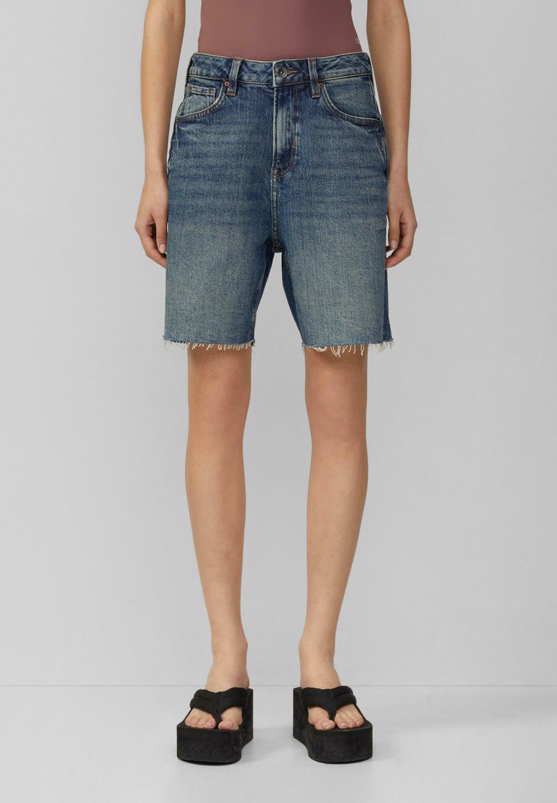 Dżinsowe szorty z wysokim stanem, postrzępionym wykończeniem i klasycznym pięciopocketowym krojem w średnim odcieniu niebieskiego. Połączone z czarnymi sandałami na platformie.