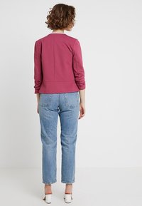 Vinröd sweatshirt med trekvartslång ärm, texturerad tyg, figursydd midja och draperad detalj, i kombination med ljusblå raka jeans.