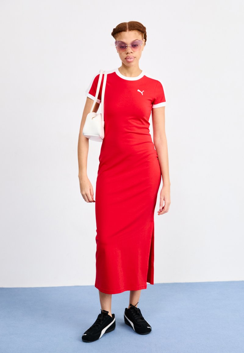 Vestido atlético rojo con mangas cortas y acentos blancos, que presenta una abertura lateral, combinado con zapatillas negras y un pequeño bolso blanco.