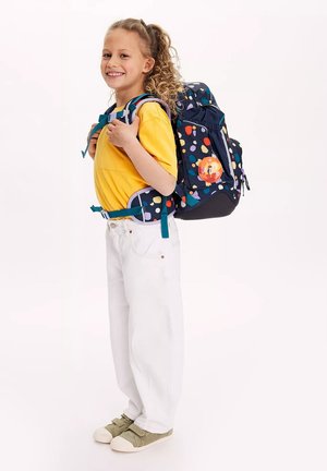 Der Rucksack hat eine marineblaue Basis mit bunten Pünktchen, verstellbaren Trägern und einem Charakter-Abzeichen. Das Kind trägt ein gelbes T-Shirt und weiße Hosen.