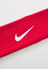 Crvena Nike traka za kosu napravljena od mekog elastičnog materijala, sa istaknutim bijelim swoosh logotipom, koja se odlikuje glatkom teksturom i ravnim dizajnom.