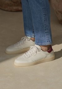 Bílé kožené tenisky s vínově červeným akcentem na patě, se zavazováním na tkaničky, perforovaným designem a krémovou gumovou podešví. Nošeny s modrými džínami.