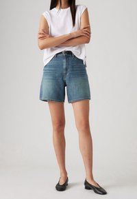 Shorts denim berwarna biru medium dengan potongan santai, pinggang tinggi, dan ujung terlipat. Dipasangkan dengan atasan lengan pendek berwarna putih dan sepatu datar hitam.