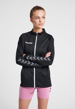 Sort zip-up sportsjakke i struktureret stof med grå chevronstriber på ærmerne og hvidt logo. Matchet med lyserøde shorts.