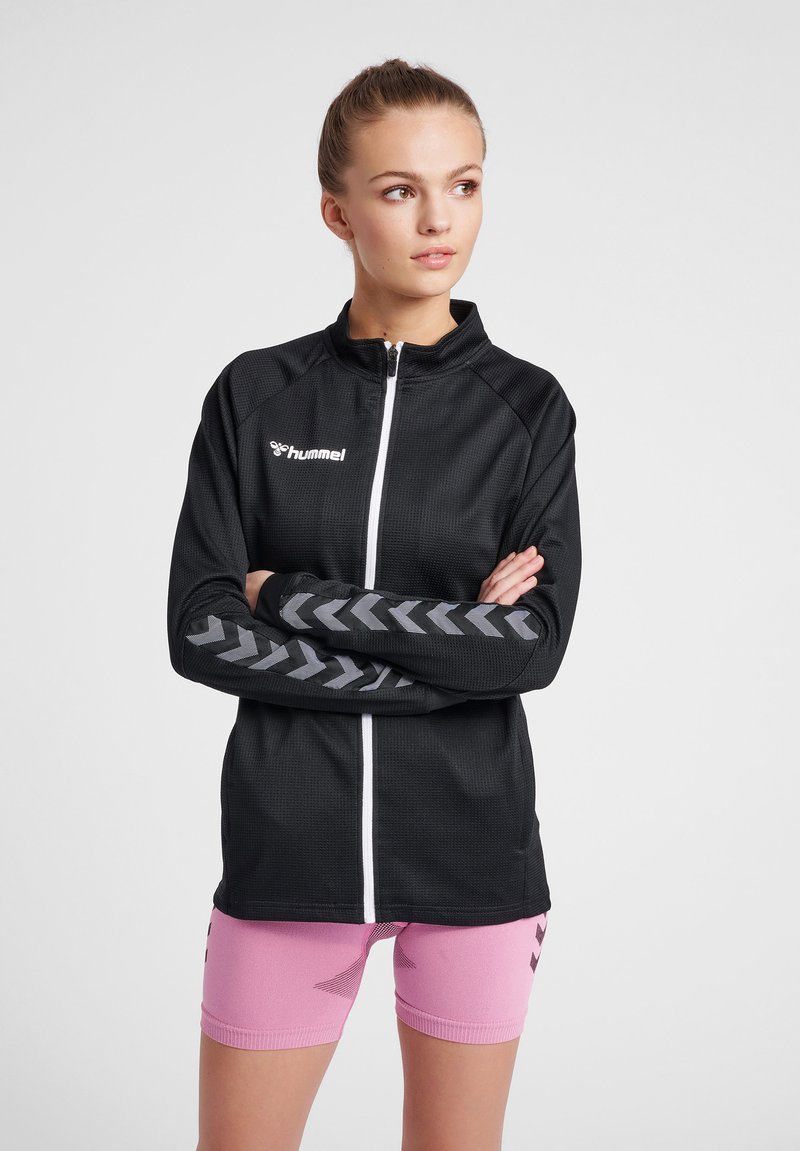 Chaqueta deportiva negra con cremallera fabricada en tejido texturizado, con rayas en forma de chevron grises en las mangas y un logo blanco. Combinada con pantalones cortos rosas.