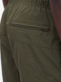 Nahaufnahme von olivgrünen Hosen mit einer geschlossenen Reißverschlusstasche auf der rechten Rückseite, getragen von einer Person mit dunkler Haut.