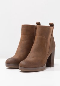 Brązowe zamszowe botki za kostkę z zaokrąglonym noskiem, bocznym zamkiem błyskawicznym, elastycznymi panelami i słupkowym obcasem. Charakteryzują się gładką teksturą i minimalnym przeszyciem.