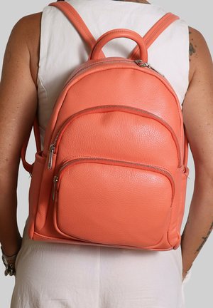 Femme portant un haut blanc sans manches avec un sac à dos en cuir corail texturé, doté de deux poches avant zippées et de bretelles rembourrées.