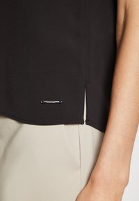 Top in tessuto nero con trama diagonale, caratterizzato da uno spacco laterale e un'accattivante accentuazione in metallo. Texture liscia, abbinato a pantaloni beige chiaro.