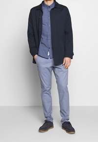 Man i en marinblå jacka, mönstrad blå skjorta med knappar, ljusblå byxor och mörka mockaskor som står mot en enkel vägg.