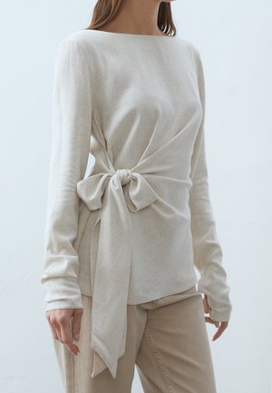 Mujer con un top beige de manga larga con un gran nudo lateral atado en la cintura, combinado con pantalones beige.
