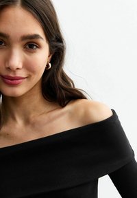 Zwarte geribbelde off-shoulder top met lange mouwen en een zachte textuur. Gouden hoepelringen accentueren de halslijn en de gezichtskenmerken.