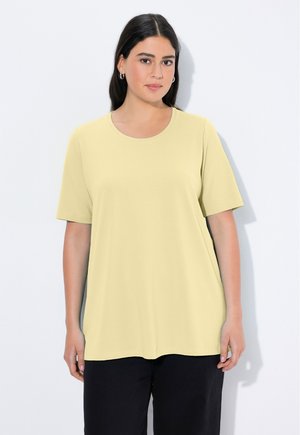 Vrouw met lang donker haar die een bleekgele korte mouw top en zwarte broek draagt, staand tegen een effen lichte achtergrond.