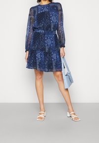 Navyblaues Kleid mit langen, transparenten Ärmeln, einem geschichteten Volantrock und Paisleymustern. Kombiniert mit einer hellblauen Handtasche und weißen Riemchensandalen.