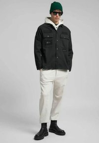 Chaqueta negra con botones y dos bolsillos en el pecho con parches de la marca. Se lleva sobre una sudadera con capucha blanca y pantalones deportivos gris claro, con botas negras. Gorro verde.