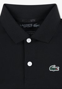 Μαύρο πόλο μπλουζάκι από υλικό με υφή, με ριγέ γιακά, που διαθέτει τρία λευκά κουμπιά και το λογότυπο κροκόδειλου Lacoste σε πράσινο και κόκκινο.